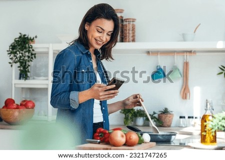 Shot of smiling beautiful woman using her mobile phone while cooking vegetables in the kitchen at home. 商業照片 © 