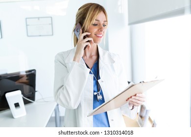 Shot Of Smiling Female Doctor Using Her Mobile Phone While Reviewing Some Papers In The Consultation.