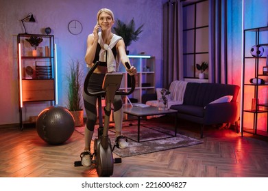 Shot of smiling fitness caucasian girl using mobile phone after training on exercise bike in evening at modern home. Portrait of model, fit sporty woman, at home engaged in a stationary bike. - Powered by Shutterstock