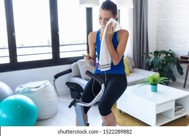 Shot of smiling fitness girl using mobile phone after training on exercise bike at home.