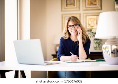 Shot Of Smiling Mature Woman Making A Phone Call And Using Laptop While Working Online. Home Office. 