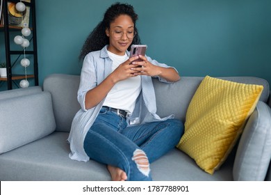 Aufnahme einer jungen, afrikanischen Amerikanerin, die ihr Handy benutzt, während sie zu Hause auf dem Sofa sitzt.