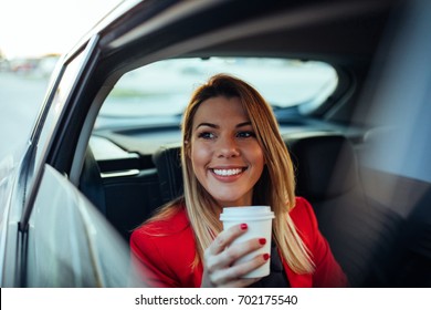 Shot Of A Smiling Young Woman Driving In The Back Seat Of The Car And Drinking Coffee.