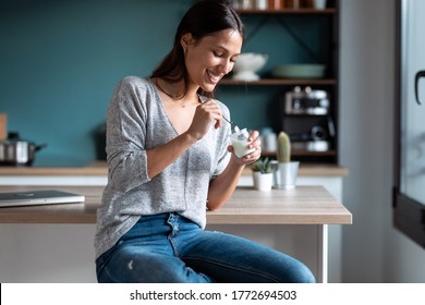 Shot of smiling young woman eating yogurt while sitting on stool in the kitchen at home.