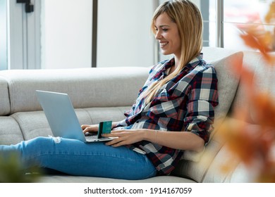 Shot Of Smiling Young Woman Paying Something Online With Her Credit Card In Laptop While Sitting On Couch At Home.