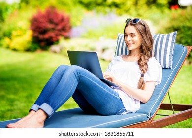Shot of smiling young woman sitting on sunbed in the garden and using her laptop.
