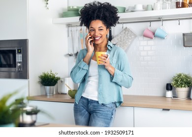 Shot Of Smiling Young Woman Talking With Her Mobile Phone While Drinking A Cup Of Coffee In The Kitchen At Home.