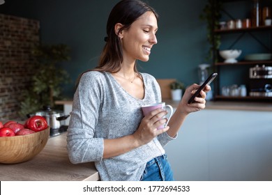 Shot of smiling young woman using her mobile phone while drinking a cup of coffee in the kitchen at home.