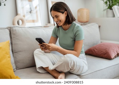 Shot of smiling young woman using her mobile phone while sitting on sofa at home