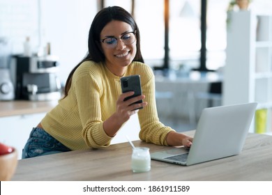 Shot of smiling young woman working with computer while sending messages with smart phone in the kitchen at home. - Powered by Shutterstock