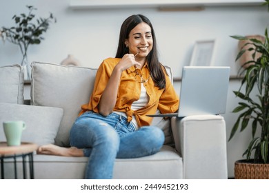 Shot of smiling young woman working with her laptop while sitting on couch at home. - Powered by Shutterstock