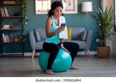 Shot Of Sporty African Young Woman Using Her Mobile Phone After Session Of Exercises While Sitting On Fitness Ball At Home.