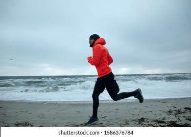 Shot Of Sporty Young Bearded Man In Black Cap, Athletic Clothes And Warm Orange Coat Running On Coastline Of Sea On Gray Stormy Weather. Sport And Healthy Lifestyle Concept