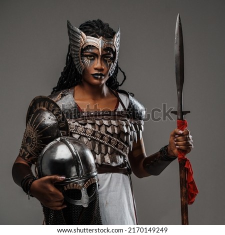 Shot of strong black woman dressed in ancient armor holding spear and helmet isolated on grey.