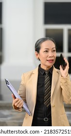 Shot Of Successful Mature Businesswoman Having Phone Conversation And Walking Outside Modern Office Building