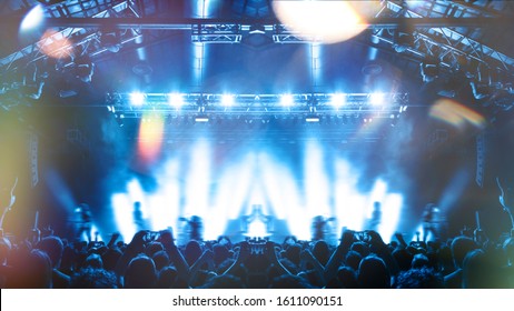 A Shot Taken In Front Of A Concert Stage Lit In The Night, People Are Visible Waving And Clapping, But No One Is Recognizable.