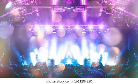 A Shot Taken In Front Of A Concert Stage Lit In The Night, People Are Visible Waving And Clapping, But No One Is Recognizable.