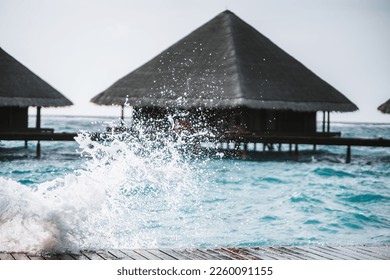 The shot is taken with a selective focus on the water splashing against a wooden footpath, leading to an overwater bungalows with an elegant design featuring thatched roofs, in the Maldives - Powered by Shutterstock