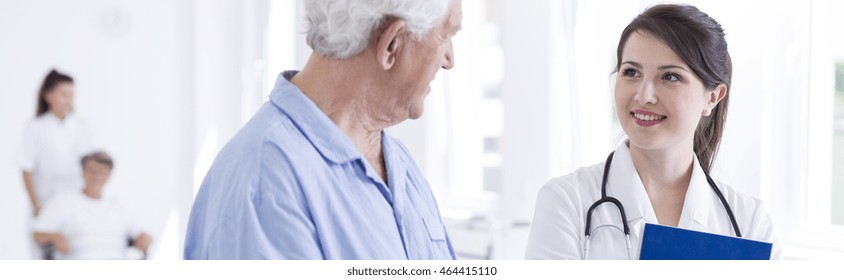 Shot Of A Talk Between Young, Smiled, Woman Doctor And The Older Patient With The Other Senior Patient With The Nurse At The Background