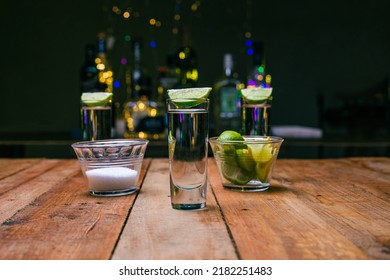 Shot Of Tequila Served In A Tequila Glass On A Wooden Table. Blurred Background. Rustic Bar Atmosphere.