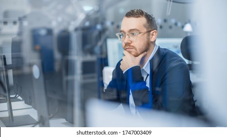 Shot Of Thoughtful Industrial Engineer Working On A Personal Computer. He Is Working In Cozy Office In Modern Factory With High-Tech Machinery.