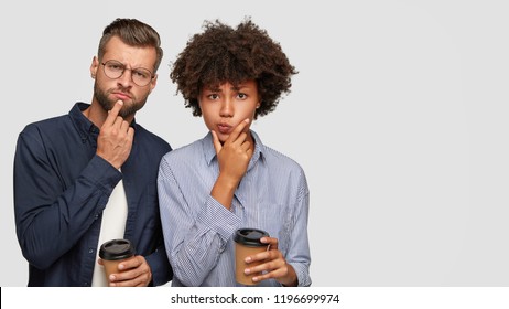 Shot of thoughtful puzzled multiethnic young couple hold chins, try to solve problem, drink aromatic takeaway coffee, stand closely against white background with copy space for your advertisement. - Powered by Shutterstock