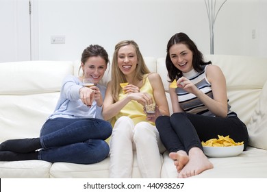 Shot Of Three Woman Sat On The Sofa At Home Watching A Film. They Are Pointing And Laughing At The TV While Eating Snacks.