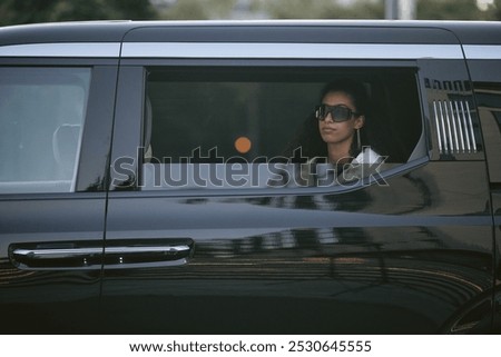 Shot through open car window of successful Middle Eastern woman in trendy sunglasses sitting in back seat of limo embodying celebrity lifestyle while arriving at award ceremony, copy space