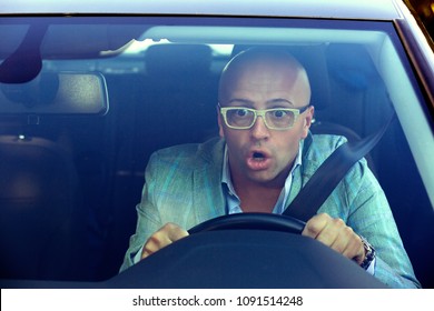Shot through windscreen of car of man in glasses driving and having accident looking extremely shocked holding steering wheel and looking away. 