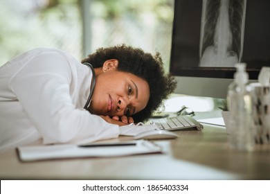 Shot Of A Tired African Female Doctor Laying Down On Her Crossed Arms Alone At Desk While Looking X-ray  Of Lungs On Computer During COVID-19 Pandemic.