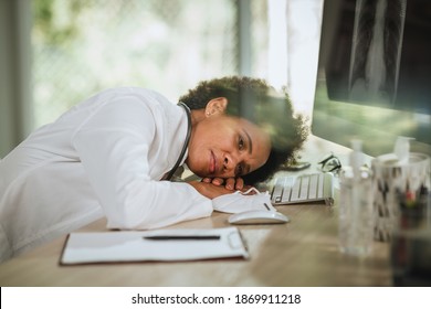 Shot Of A Tired African Female Doctor Laying Down On Her Crossed Arms Alone At Desk While Looking X-ray On Computer During COVID-19 Pandemic.