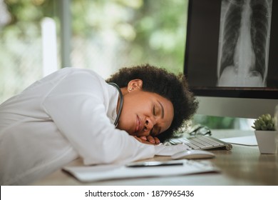 Shot Of A Tired African Female Doctor Laying Down On Her Crossed Arms At Desk While Looking X-ray  Of Lungs On Computer During COVID-19 Pandemic.