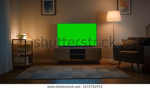 Shot of a\
TV with Horizontal Green Screen Mock Up. Cozy Evening Living Room\
with a Chair and Lamps Turned On at\
Home.