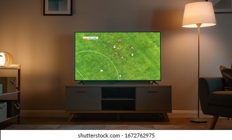 Shot Of A TV With Soccer Match. Cozy Evening Living Room With A Chair And Lamps Turned On At Home.