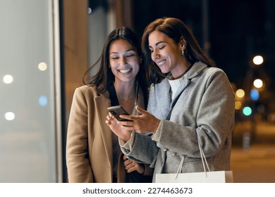 Shot of two beautiful friends talking and using smartphone while walking on the city street with shopping bag.