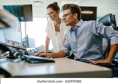 Shot Of Two Business Colleagues Working In Their Office Using A Desktop Computer. Man Sitting At His Desk With Female Colleague Standing By.