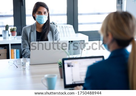 Shot of two business women wearing a hygienic face mask while work with laptops in the coworking space. Social distancing concept.