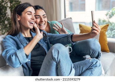 Shot Of Two Happy Beautiful Women Doing A Video Call With Laptop Sitting On The Couch At Home.