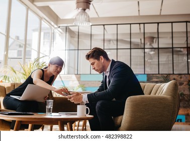Shot Of Two People Sitting In Office Lobby. Business Partners Working Together In Modern Office Looking At Mobile Phone.