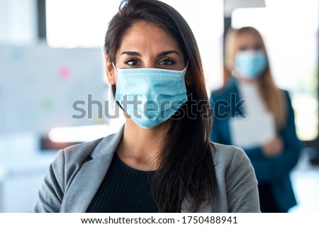 Shot of two pretty business women wearing a hygienic face mask while looking at camera in the coworking space. Social distancing concept.