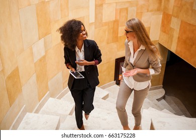 Shot Of Two Smiled Businesswomen Going Upstairs. Women Wearing Suits. One Woman Is Black.