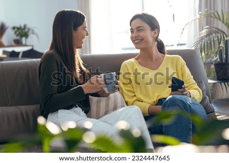 Shot of two smiling young women talking while drinking coffee sitting on couch in the living room at home.