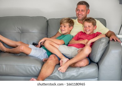 A shot of two young brother's sitting down on a couch together watching tv with their dad, they are wearing casual clothing.