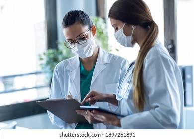 Shot Of Two Young Women Doctor Wearing A Hygienic Facial Mask Talking With Her Colleague While Working Digital Tablet In Hospital.