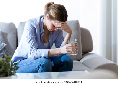 Shot of unhealthy young woman with headache holding glass of water while sitting on sofa at home.