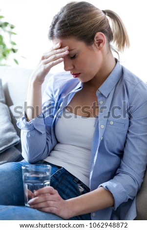 Shot of unhealthy young woman with headache holding glass of water while sitting on sofa at home.