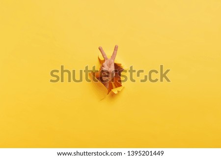 Shot of unrecognizable man demonstrates victroy sign through torn hole in yellow paper, expresses celebration and triumph. Unusual concept. Copy space. Breaking through paper wall. Body language