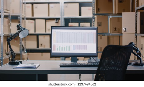 Shot of a Warehouse Inventory Manager Desk and Personal Computer wth Opened Spreadsheet. In the Background Shelves Full of Cardboard Box Packages Ready For Shipping.