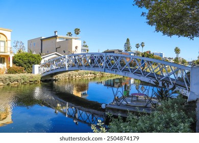 a shot of a white wooden bridge over the canal water with lush green trees and plants reflecting off the water with blue sky at the Venice Canals in Venice, Los Angeles California - Powered by Shutterstock