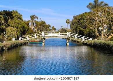 a shot of a white wooden bridge over the canal water with lush green trees and plants reflecting off the water with blue sky at the Venice Canals in Venice, Los Angeles California - Powered by Shutterstock